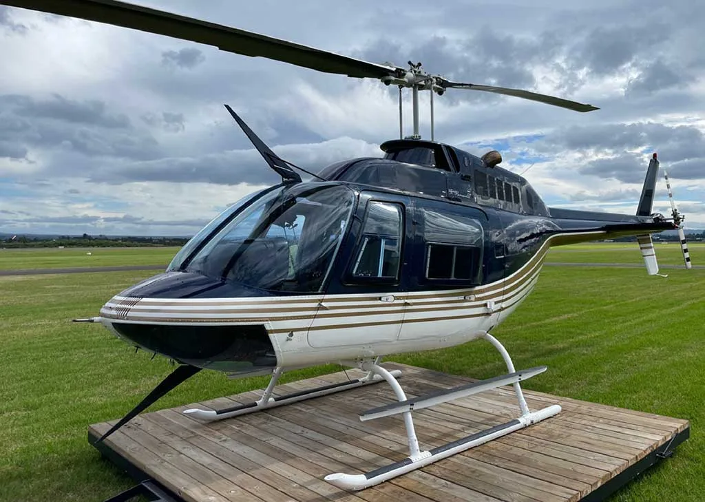 A black and white helicopter with gold stripes sits on a wooden platform on a grassy field under a cloudy sky.