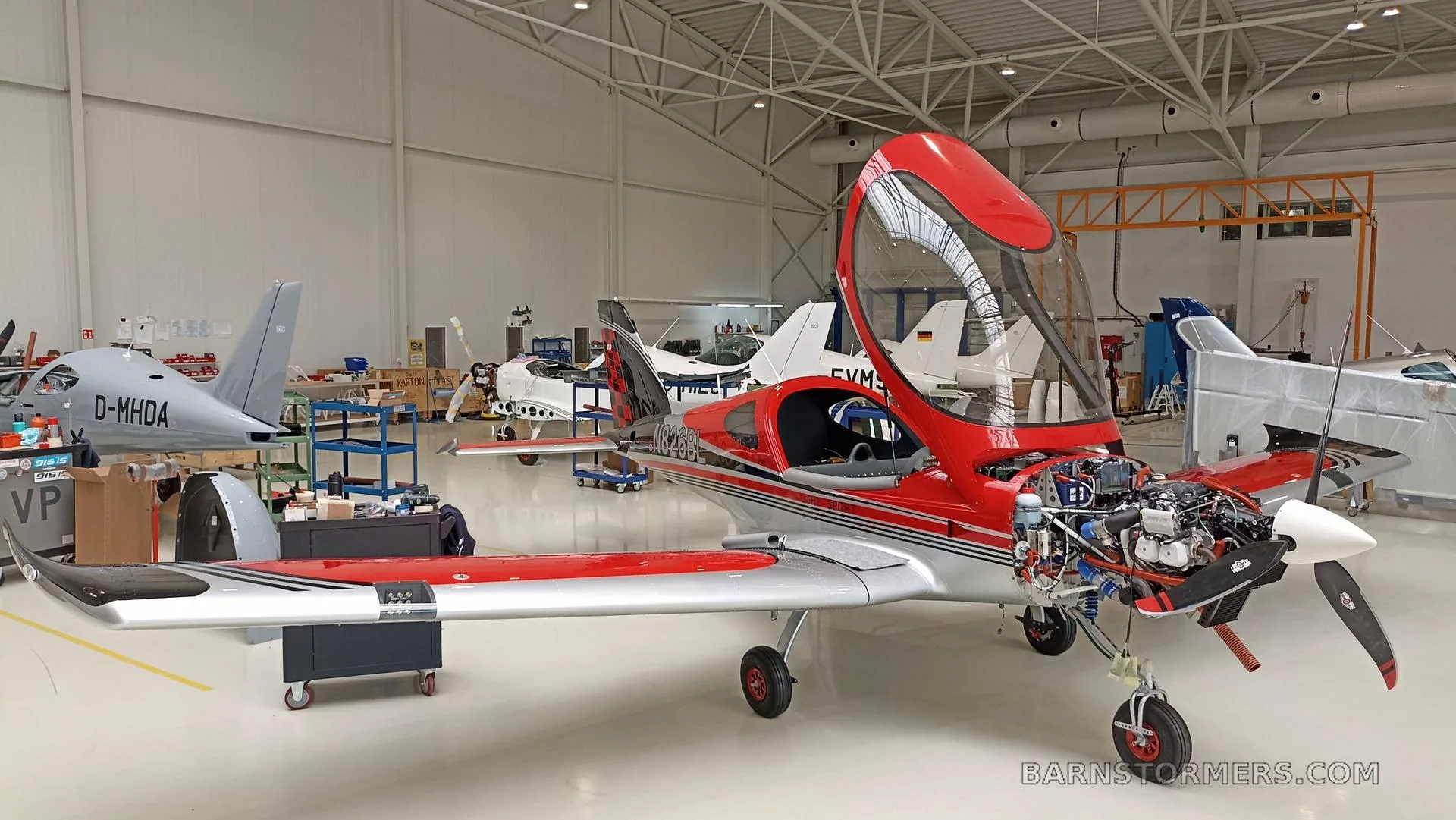 A red and silver Bristell aircraft with its engine cowling open for maintenance, parked in a large hangar with other aircraft.