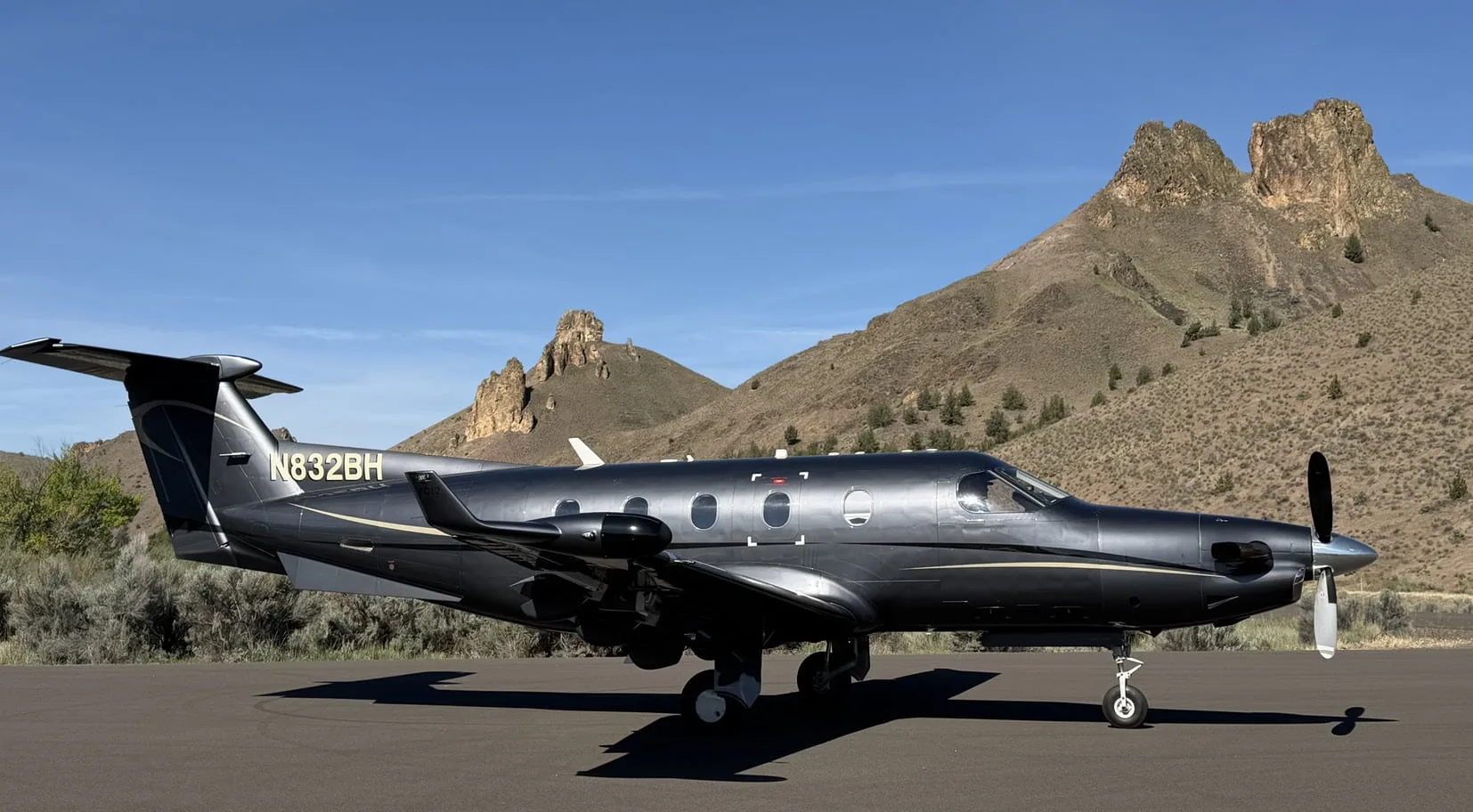A dark grey Pilatus PC-12 aircraft with gold accents, registration N832BH, parked on an airfield with rugged mountains in the background under a clear blue sky.