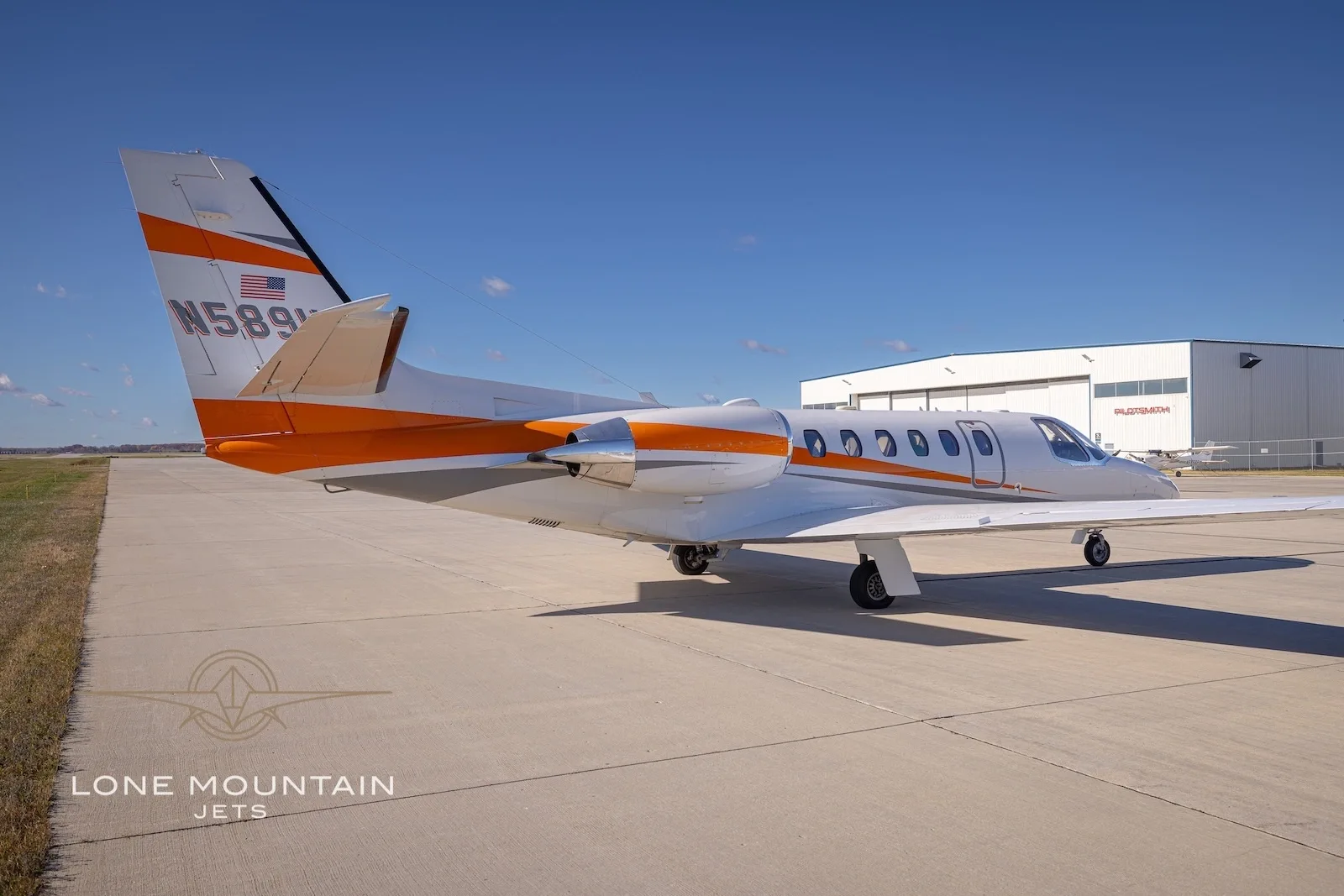 A white Cessna Citation Bravo business jet with orange and grey stripes parked on the tarmac with a hangar in the background.