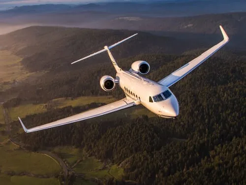 A white private jet, possibly a Gulfstream, flying over a lush green, forested landscape at sunset.