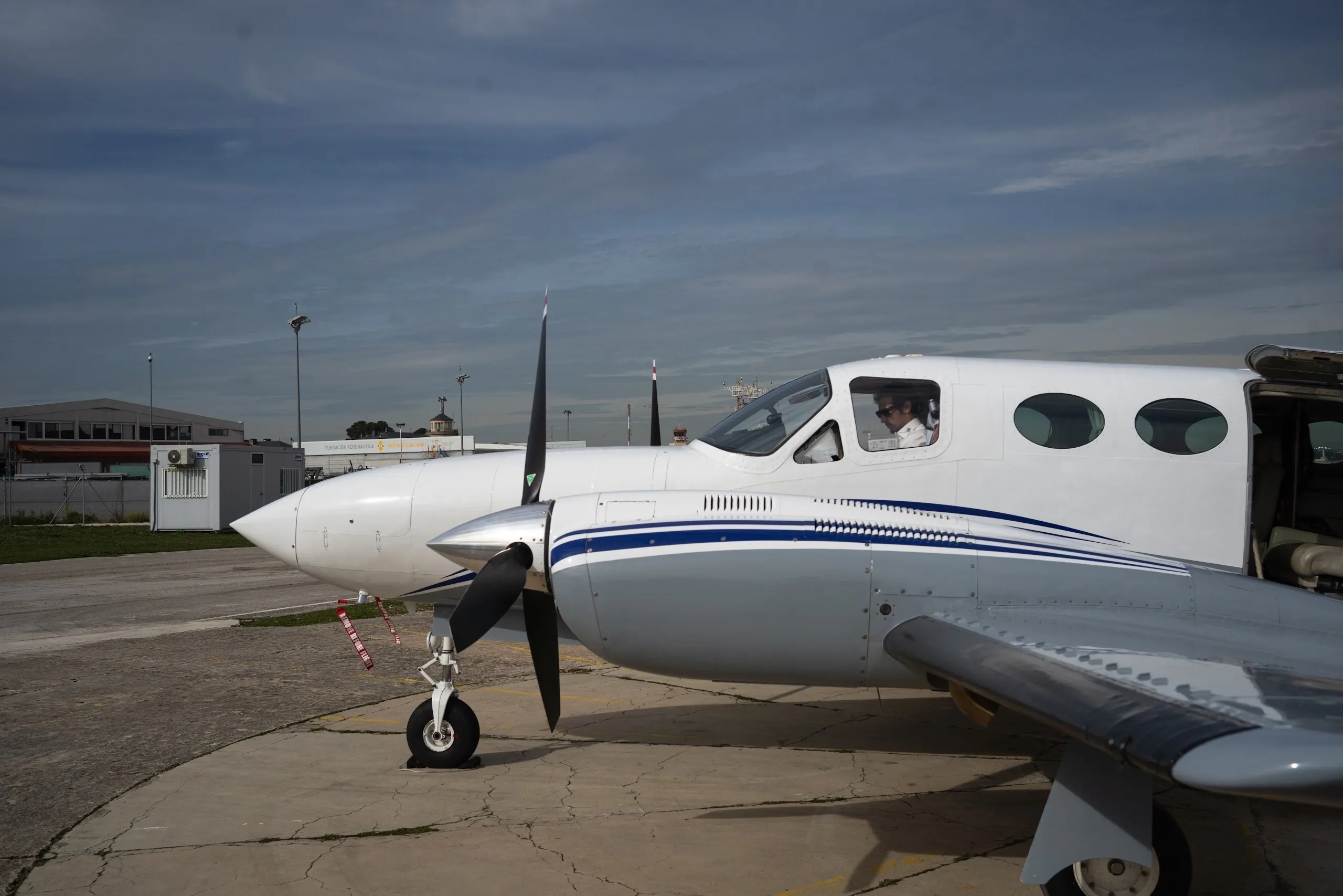 A side view of a white and grey twin-engine aircraft with blue stripes, showing a pilot in the cockpit and the passenger door open.