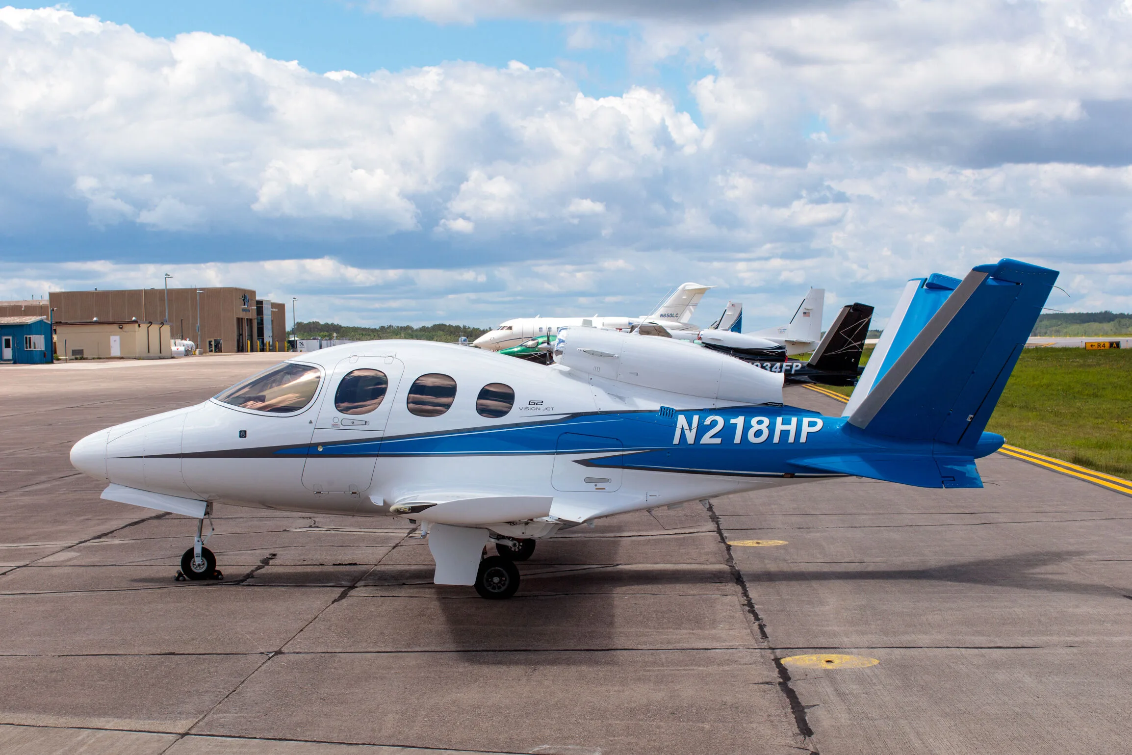 A white and blue Cirrus Vision Jet (N218HP) parked on an airport tarmac with other jets in the background.