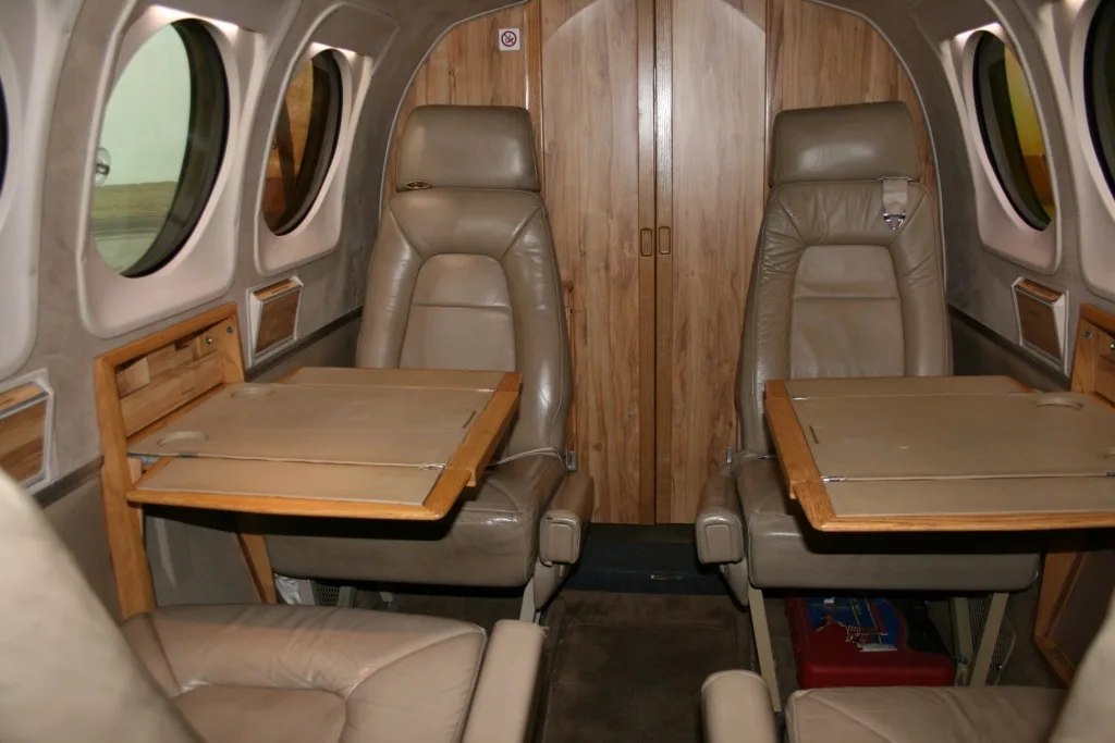 The aircraft's interior cabin featuring two tan leather seats with fold-out tables and wood paneling.