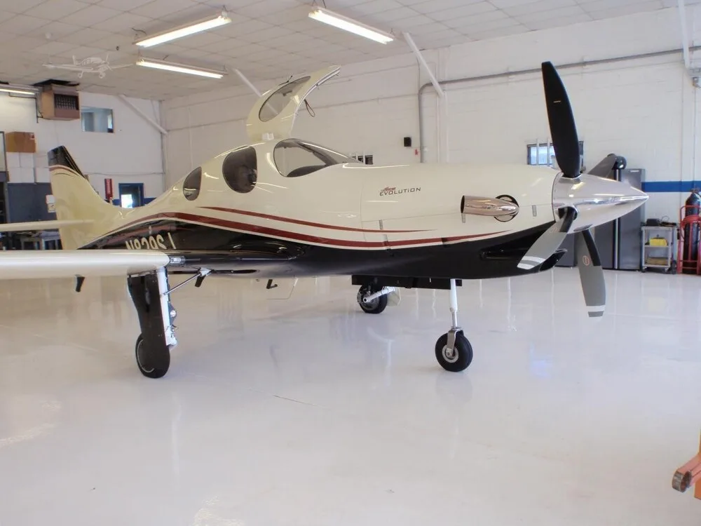 A white and black Lancair Evolution aircraft with red stripes, parked in a hangar with its doors open.