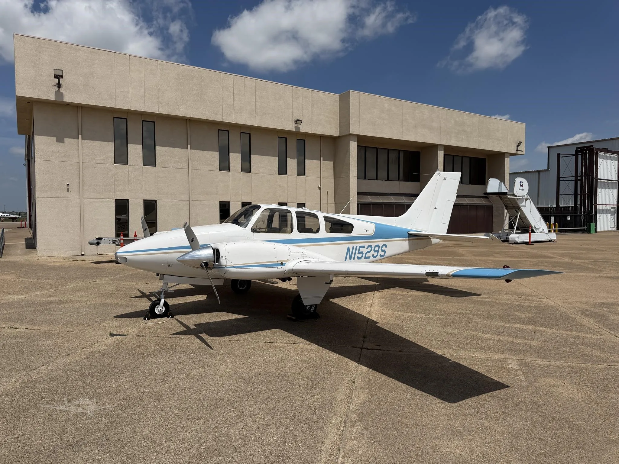 A white and blue twin-engine propeller aircraft, N1529S, parked on an apron in front of a beige building under a partly cloudy sky.