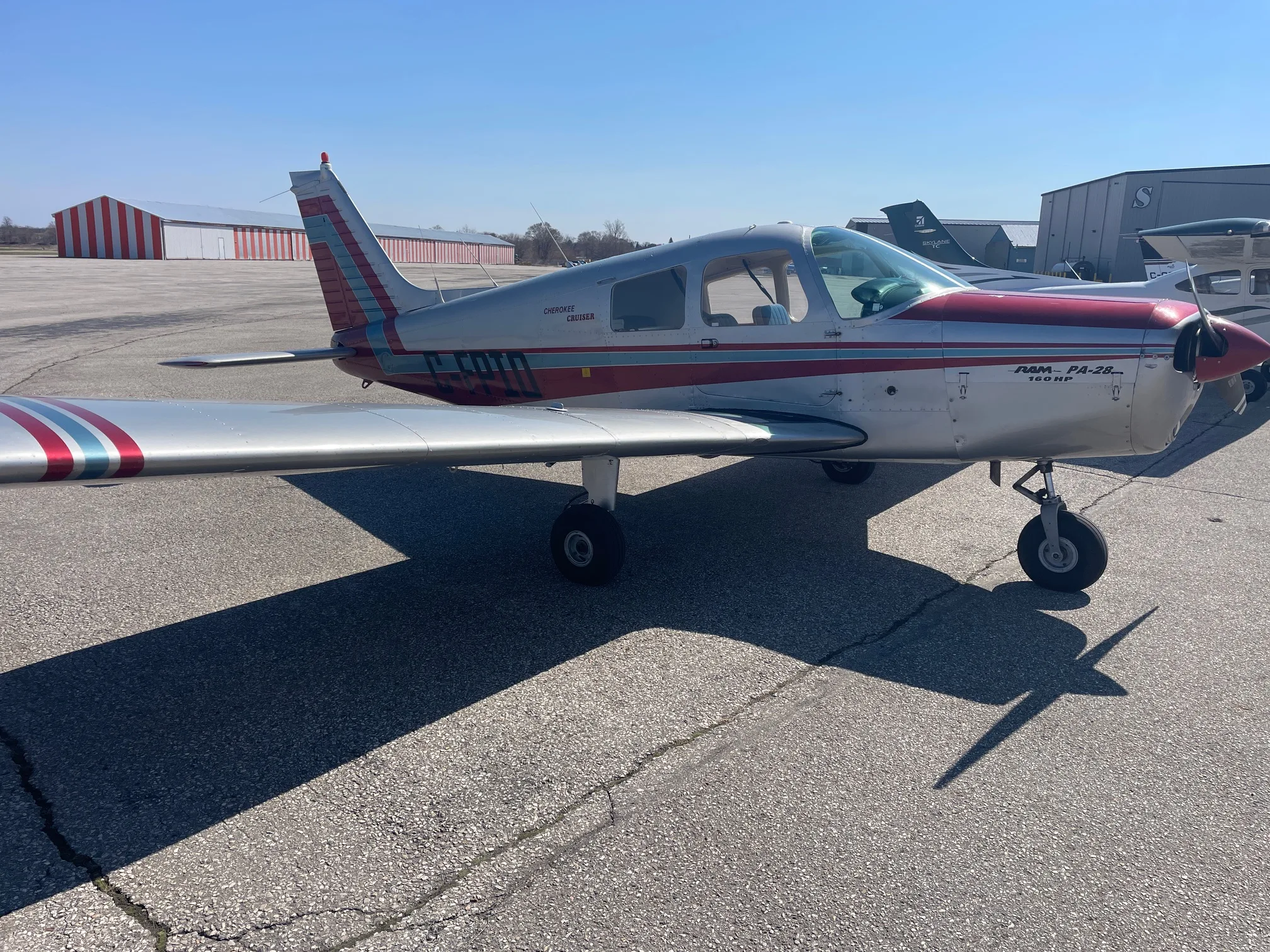 A silver and red Piper PA-28 Cherokee Cruiser aircraft parked on an airfield with hangars in the background.
