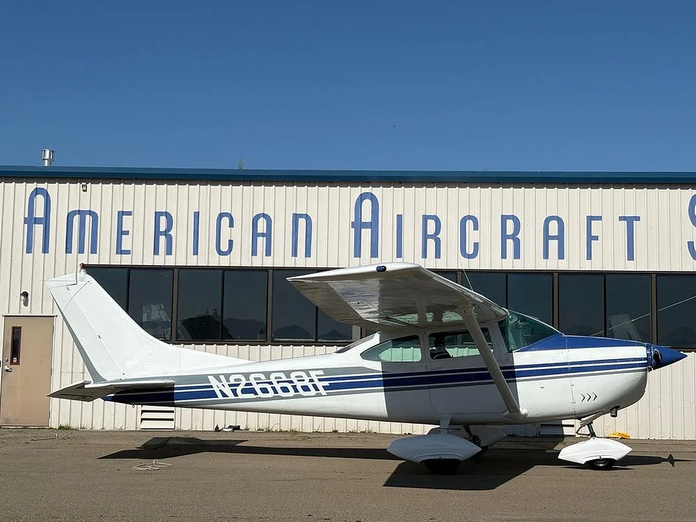 A white and blue Cessna 182 aircraft (N2668F) parked in front of an "American Aircraft Sales" hangar, viewed from the rear-right.