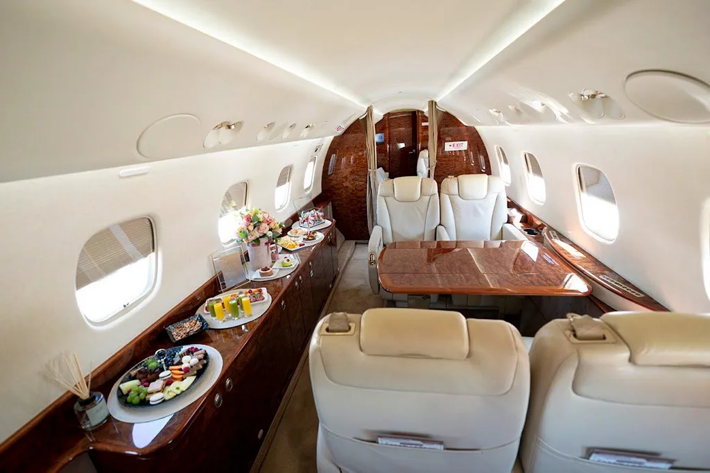 A view of the business jet cabin showing beige leather seats, a dining table, and a side counter laden with snacks and beverages.