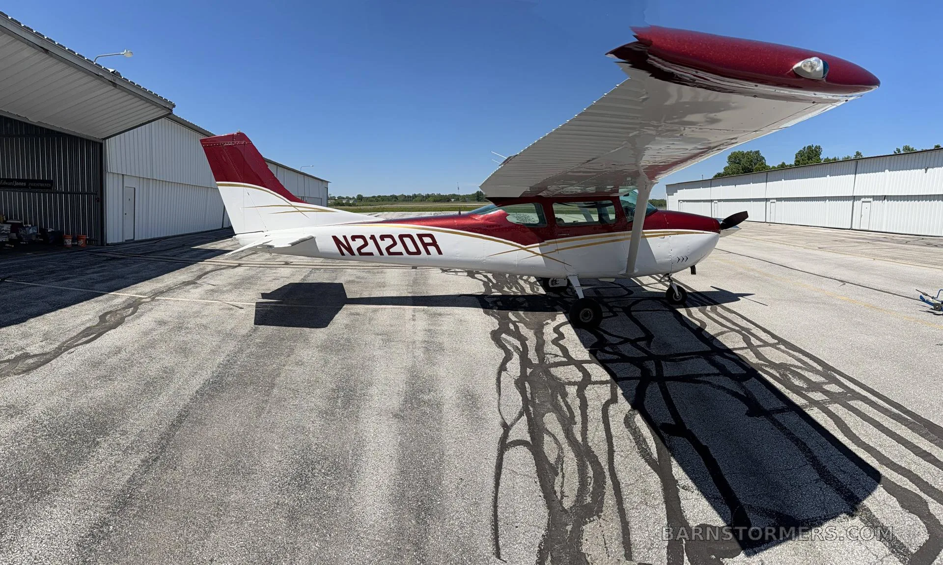 A full side view of a red and white Cessna 182 aircraft with tail number N2120R parked on an airport tarmac next to a hangar.