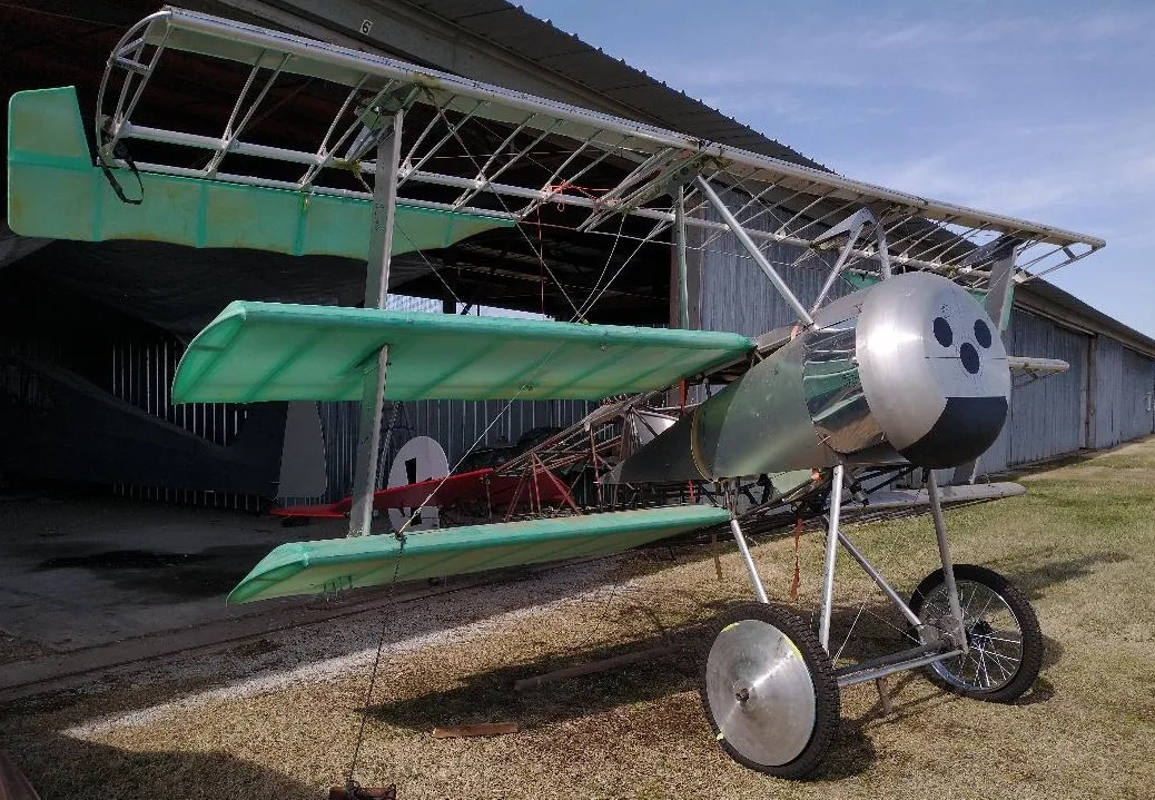 A silver-fuselaged triplane with translucent green wings parked outside a hangar.