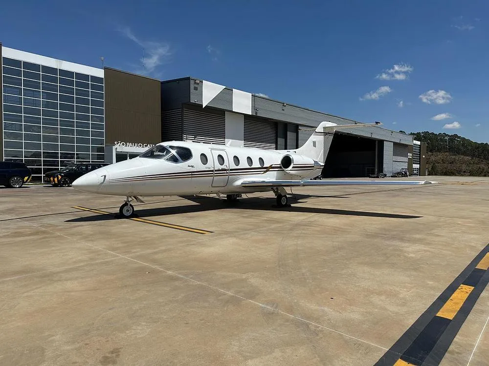 A white business jet with brown stripes parked on a tarmac in front of a modern hangar under a blue sky.