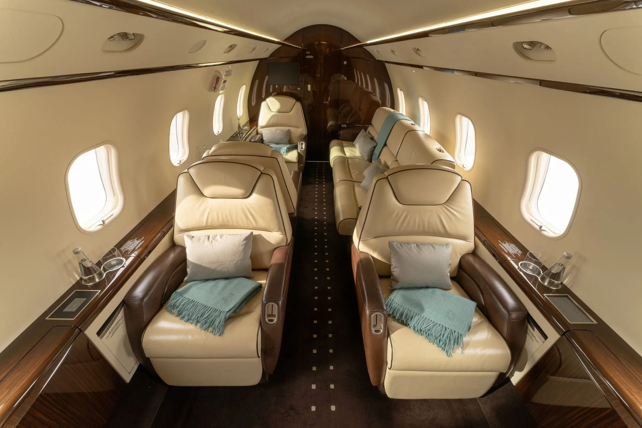 The aircraft cabin shows luxurious beige leather seats with teal throws and pillows, set against dark wood paneling.