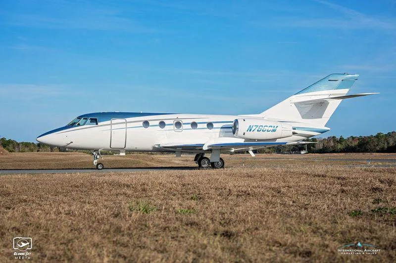A white and blue business jet N786CM is parked on a grassy field under a clear blue sky.