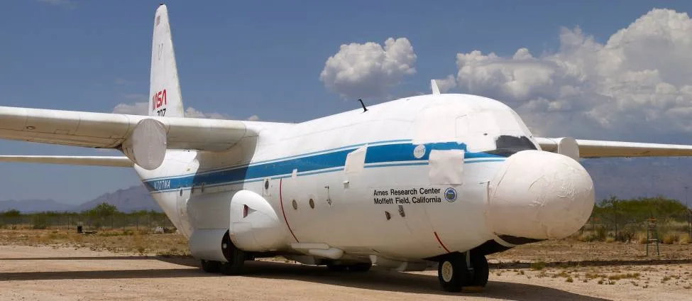 A white NASA C-130 aircraft with covered nose and engine inlets is parked in a desert boneyard under a partly cloudy sky.