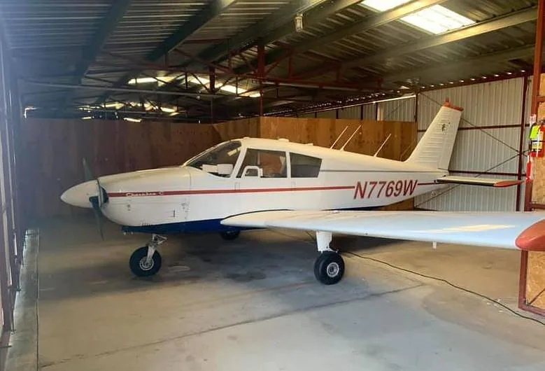 A white Piper Cherokee C aircraft with red and blue stripes, registration N7769W, parked inside a hangar.