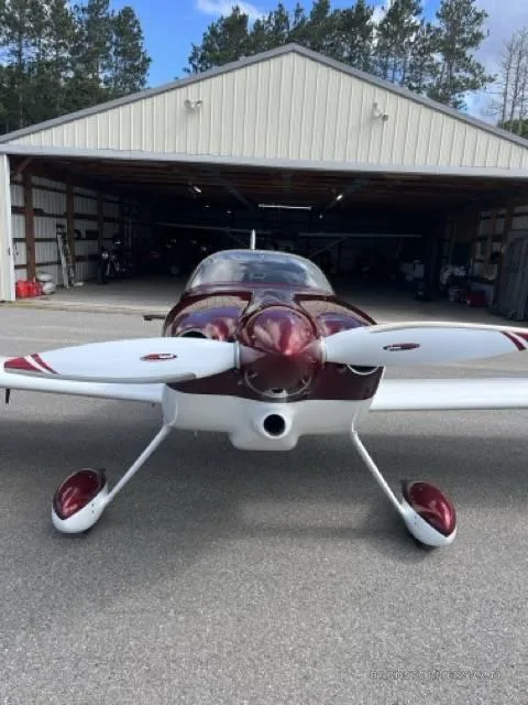 A front view of a red and white single-engine aircraft with a propeller, parked on an asphalt apron in front of a hangar.