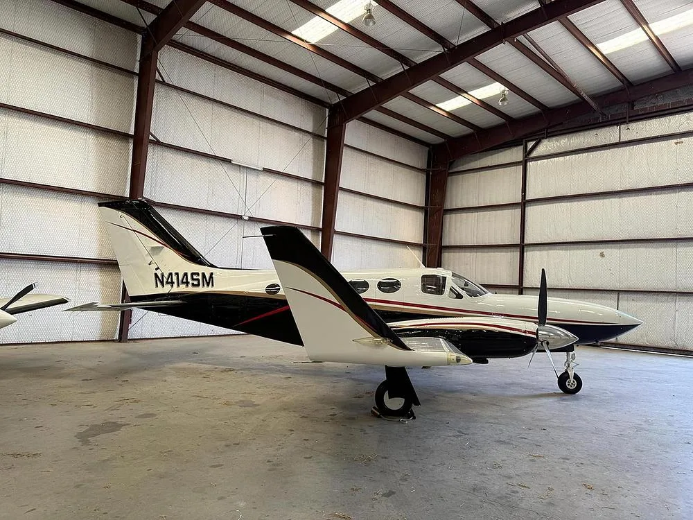 A white and black twin-engine aircraft, N414SM, with gold trim, parked inside a hangar, viewed from the rear-right.