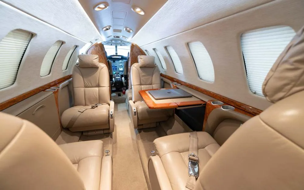The interior of a private jet cabin featuring beige leather seats, a fold-out table with a laptop, and wood accents.