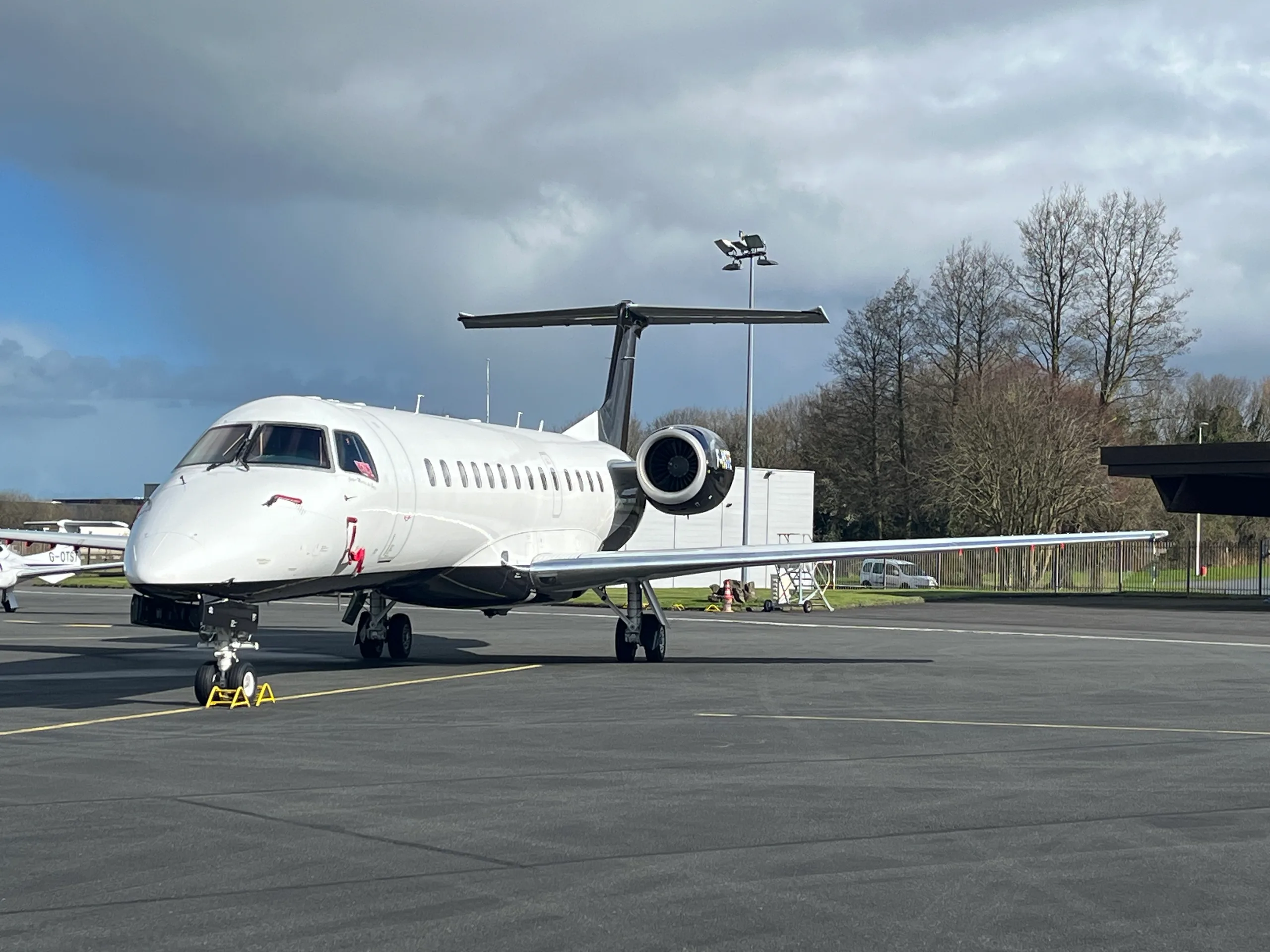 A white business jet with dark engines and tail, parked on an asphalt tarmac under a partly cloudy sky.