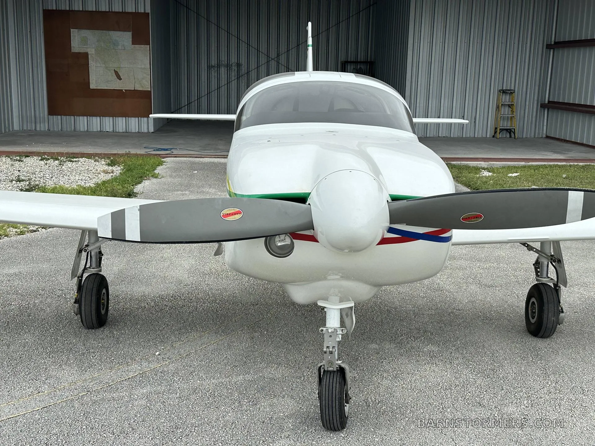 Front view of a white single-engine propeller aircraft with green and red/blue stripes, parked in front of a hangar.