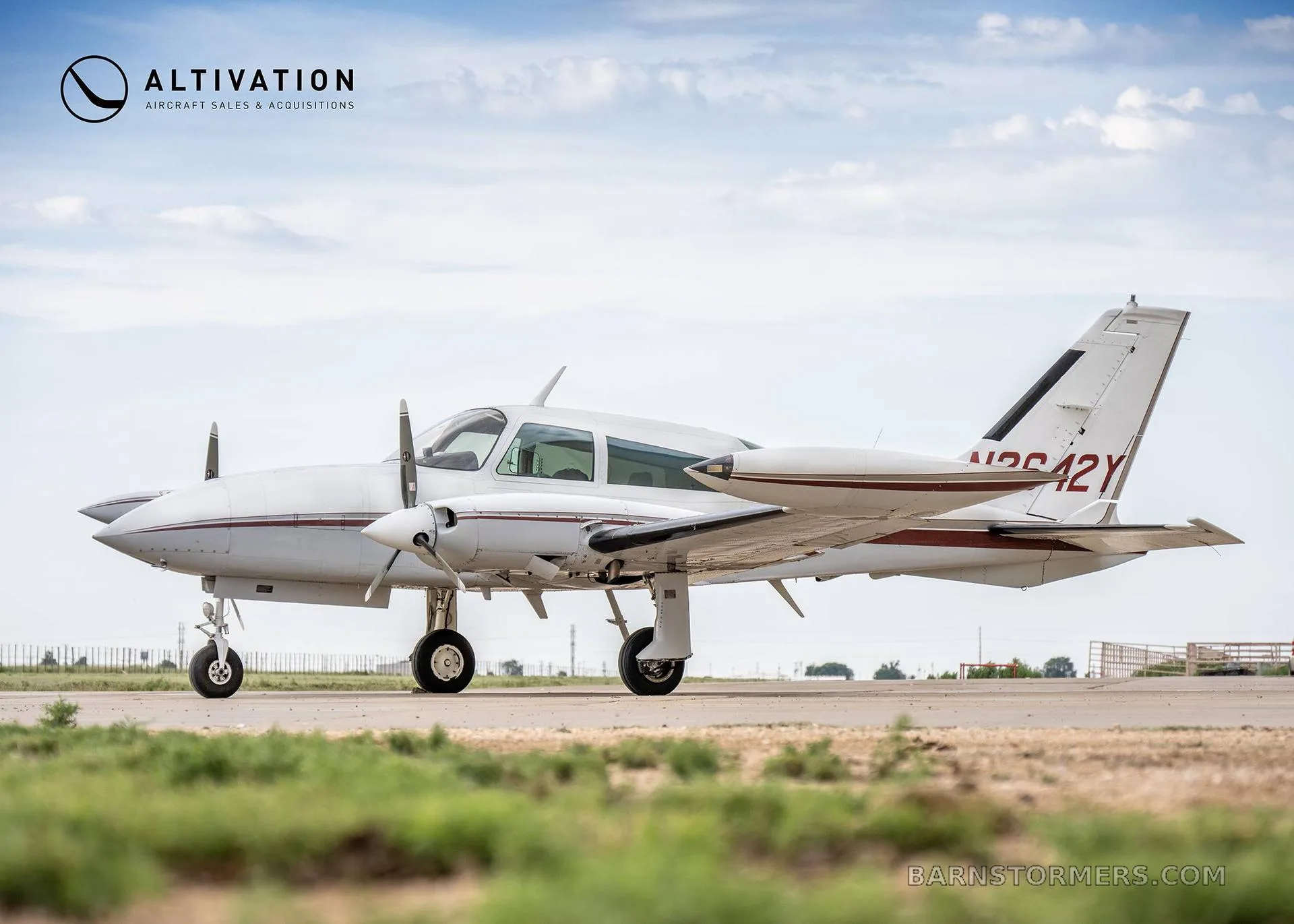 A white twin-engine propeller aircraft with maroon stripes, registration N2642Y, parked on an asphalt runway under a partly cloudy sky.