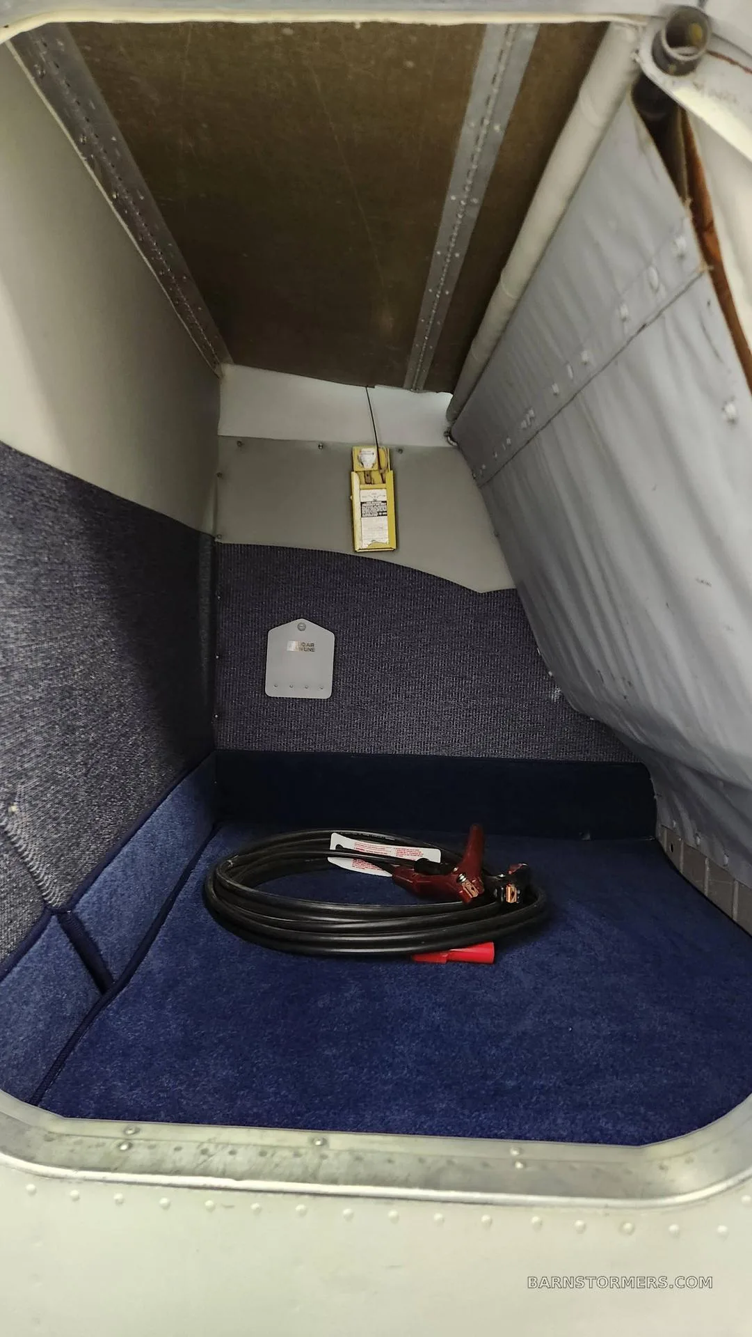 An interior view of an aircraft's baggage compartment, showing blue carpeted walls and floor with a coiled cable and jumper clamps.