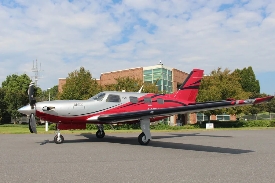 A Piper M600 aircraft, registration N501RL, in a red and silver paint scheme, parked on a tarmac with a building in the background.