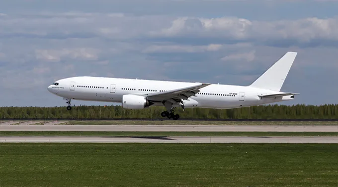 A large, all-white Boeing 777 aircraft with landing gear extended, flying low over a runway with green grass.