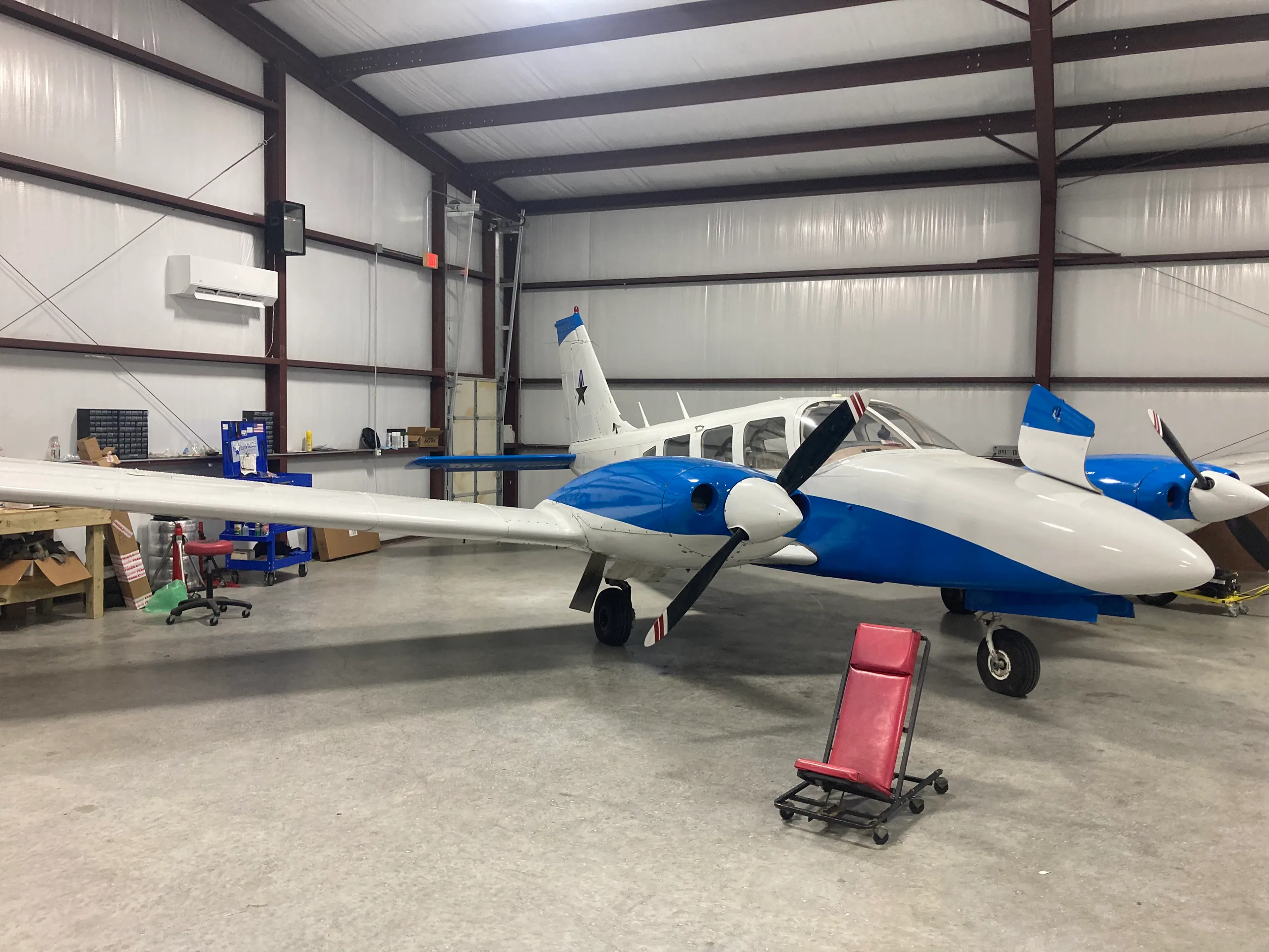 A white and blue twin-engine propeller aircraft, likely a Piper Seneca, is parked in a hangar.