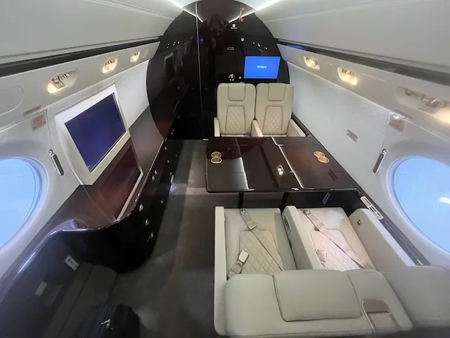 A wide view of the business jet cabin interior, showing a dining area, lounge sofa, and credenza with a screen.