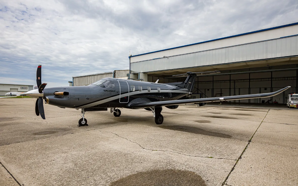 A dark grey and black Pilatus PC-12 turboprop aircraft with a silver stripe is parked on an asphalt apron in front of a hangar under a cloudy sky.