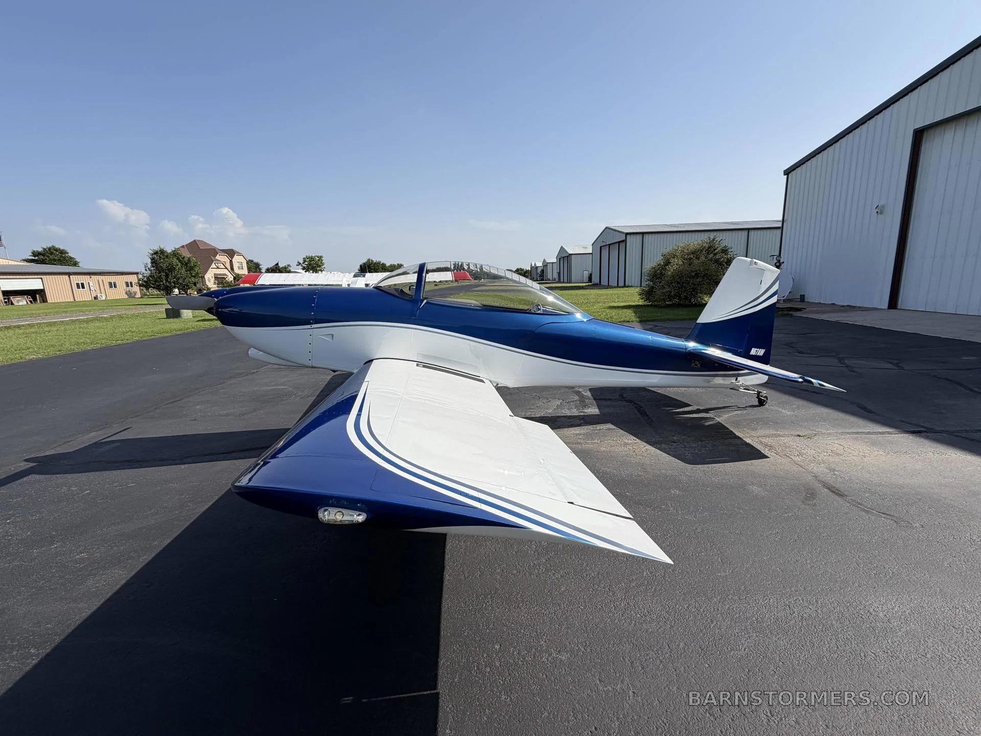 A blue and white single-engine aircraft is parked on a tarmac next to hangars under a clear blue sky.