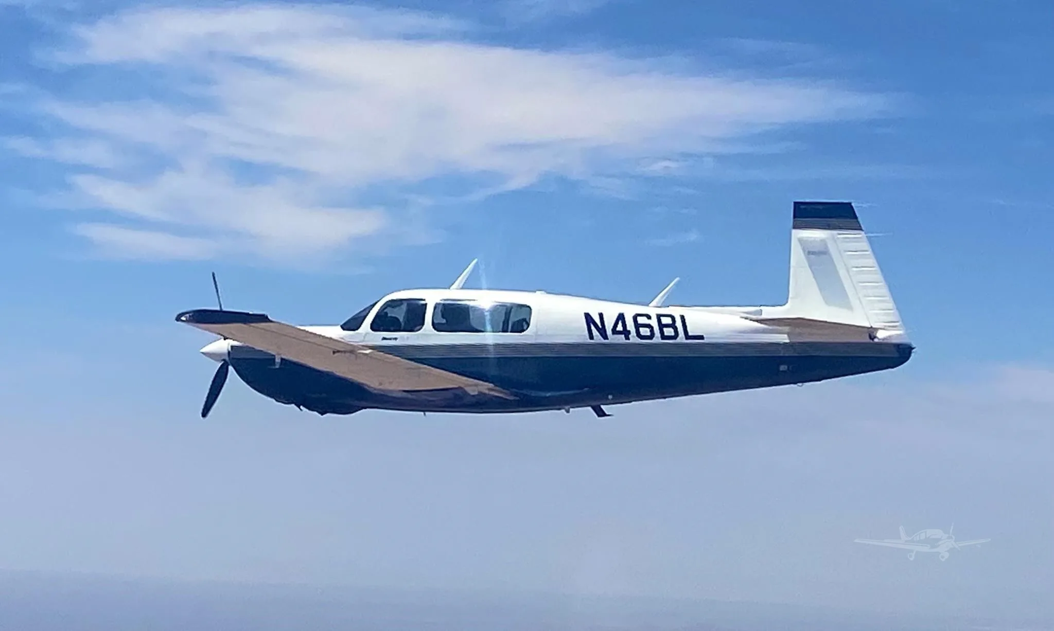 A white and dark blue single-engine propeller aircraft, registration N46BL, flying in a blue sky with scattered clouds.