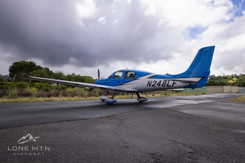 A rear-side view of a blue and silver single-engine propeller aircraft (Cirrus SR22) with registration N248LF parked on a wet tarmac under a cloudy sky.