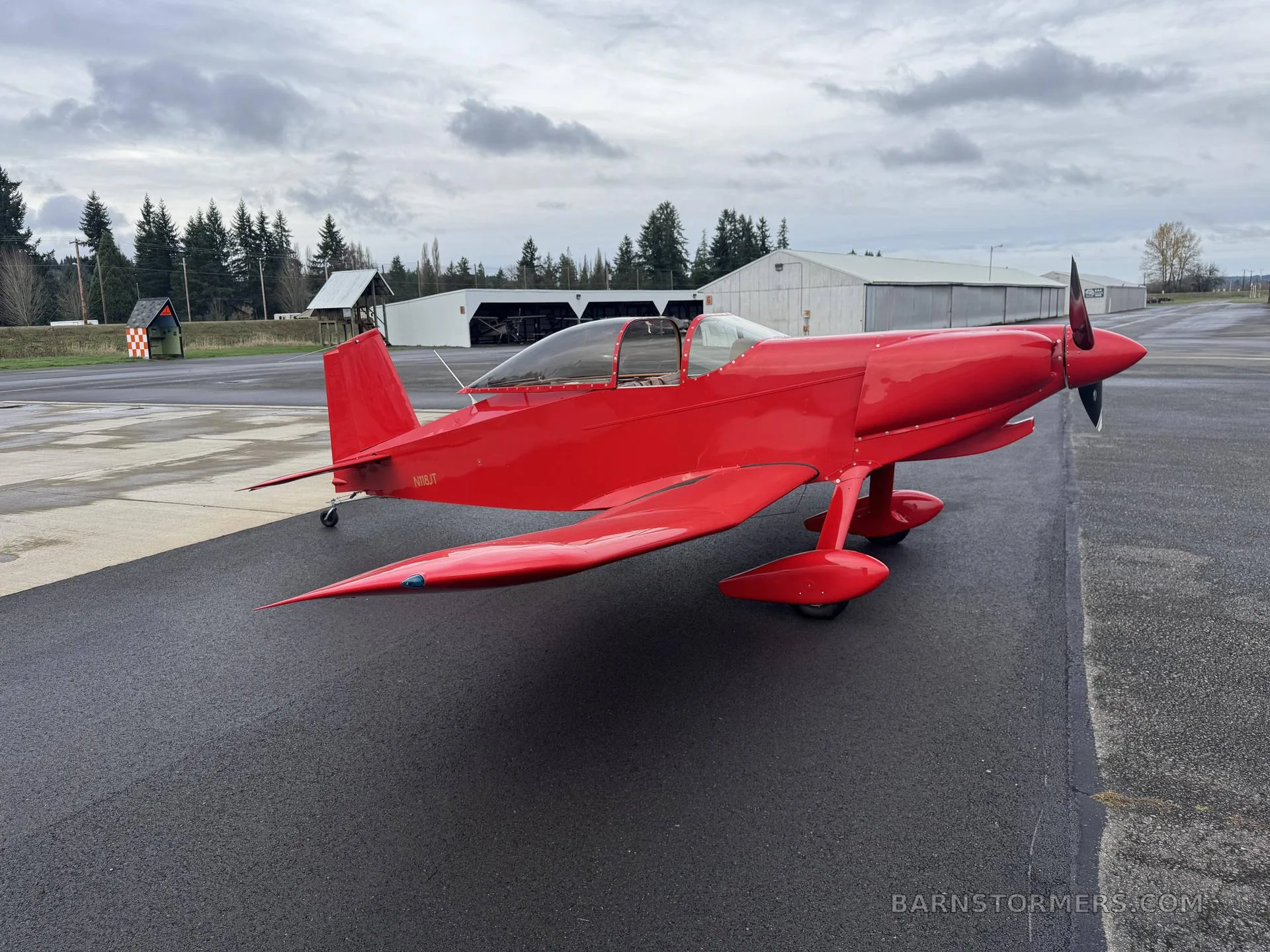 A red single-engine experimental aircraft (N18JT) with a bubble canopy is parked on an asphalt tarmac under a cloudy sky.