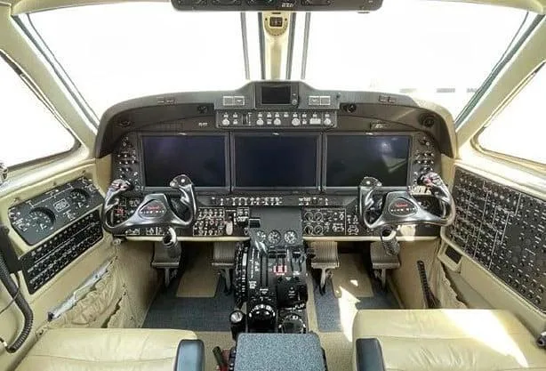 The cockpit of an aircraft featuring three large display screens, dual control yokes, and a central throttle quadrant.