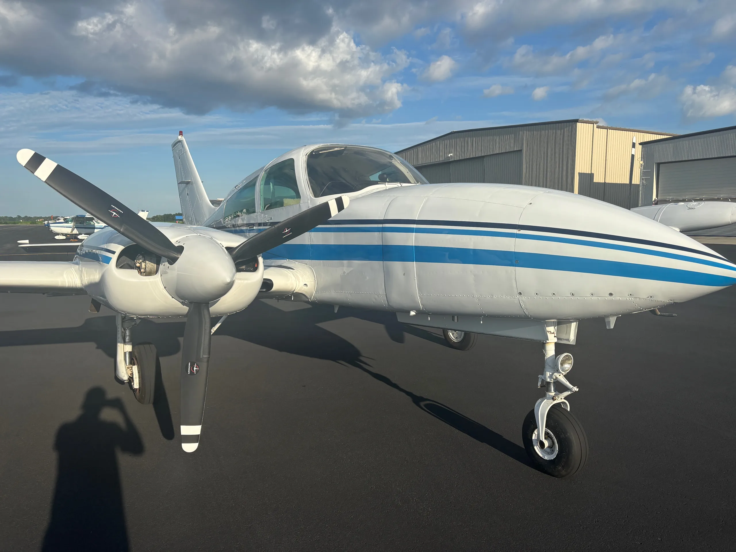 A white twin-engine aircraft with blue stripes on the tarmac, with hangars in the background and a person's shadow.