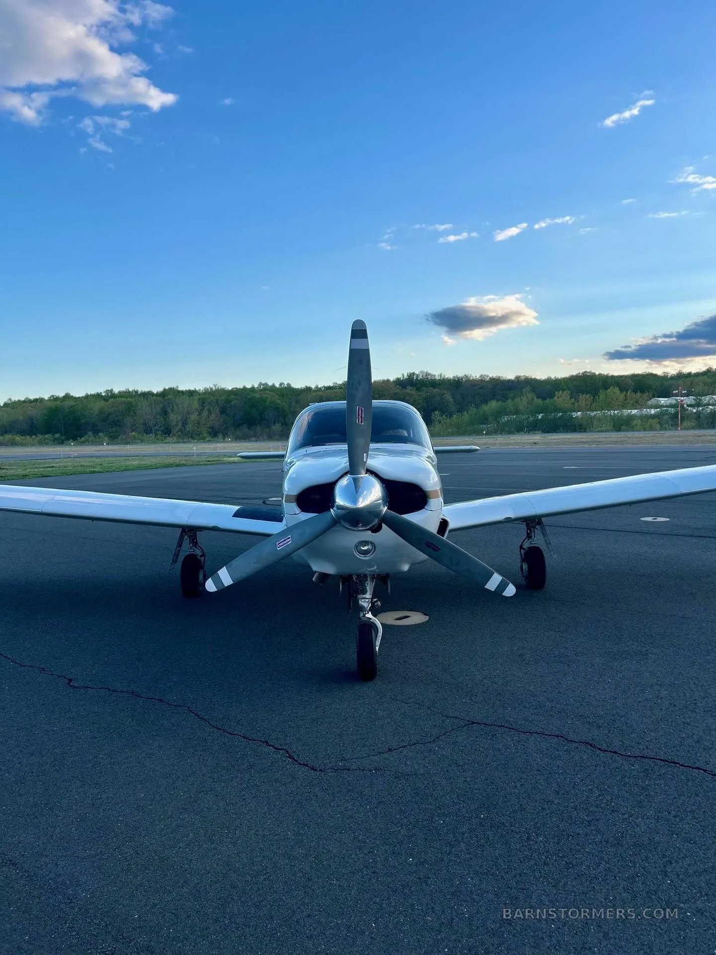 A white single-engine propeller aircraft, viewed from the front, on an asphalt tarmac under a blue sky with clouds.