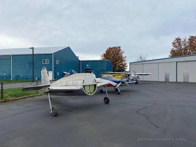 A bare metal aircraft fuselage, partially assembled, is parked on an airfield next to other small planes and blue hangars.