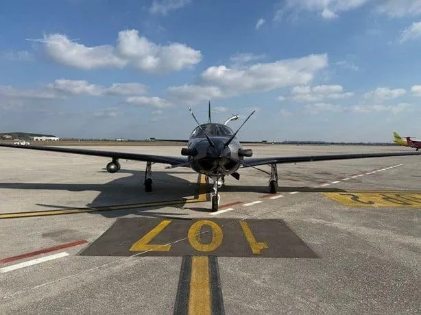 A dark grey Piper M600/SLS turboprop aircraft viewed from the front, parked on an airport tarmac with runway markings.