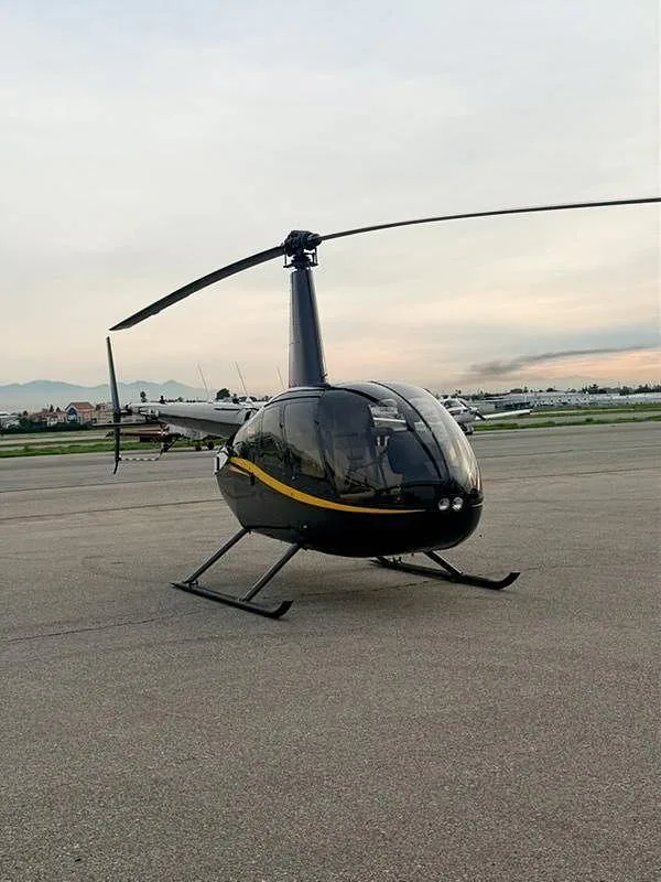 A black Robinson R44 helicopter with a yellow stripe, parked on a wide asphalt tarmac at sunset.