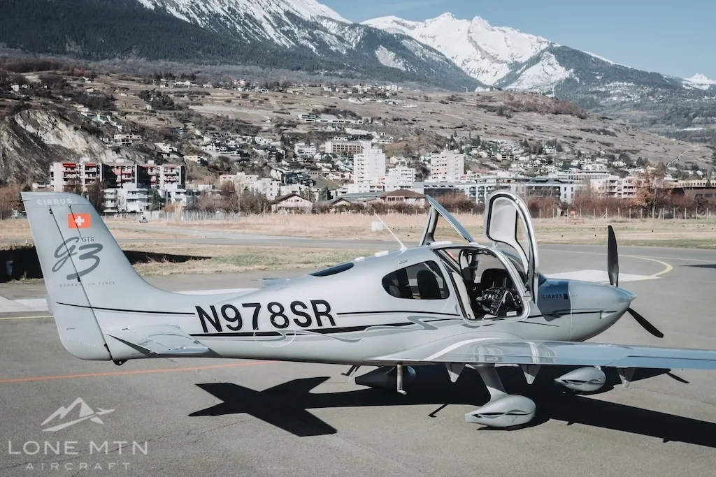 A silver Cirrus SR22 aircraft (N978SR) with its doors open, parked on an airfield with mountains and a town in the background.