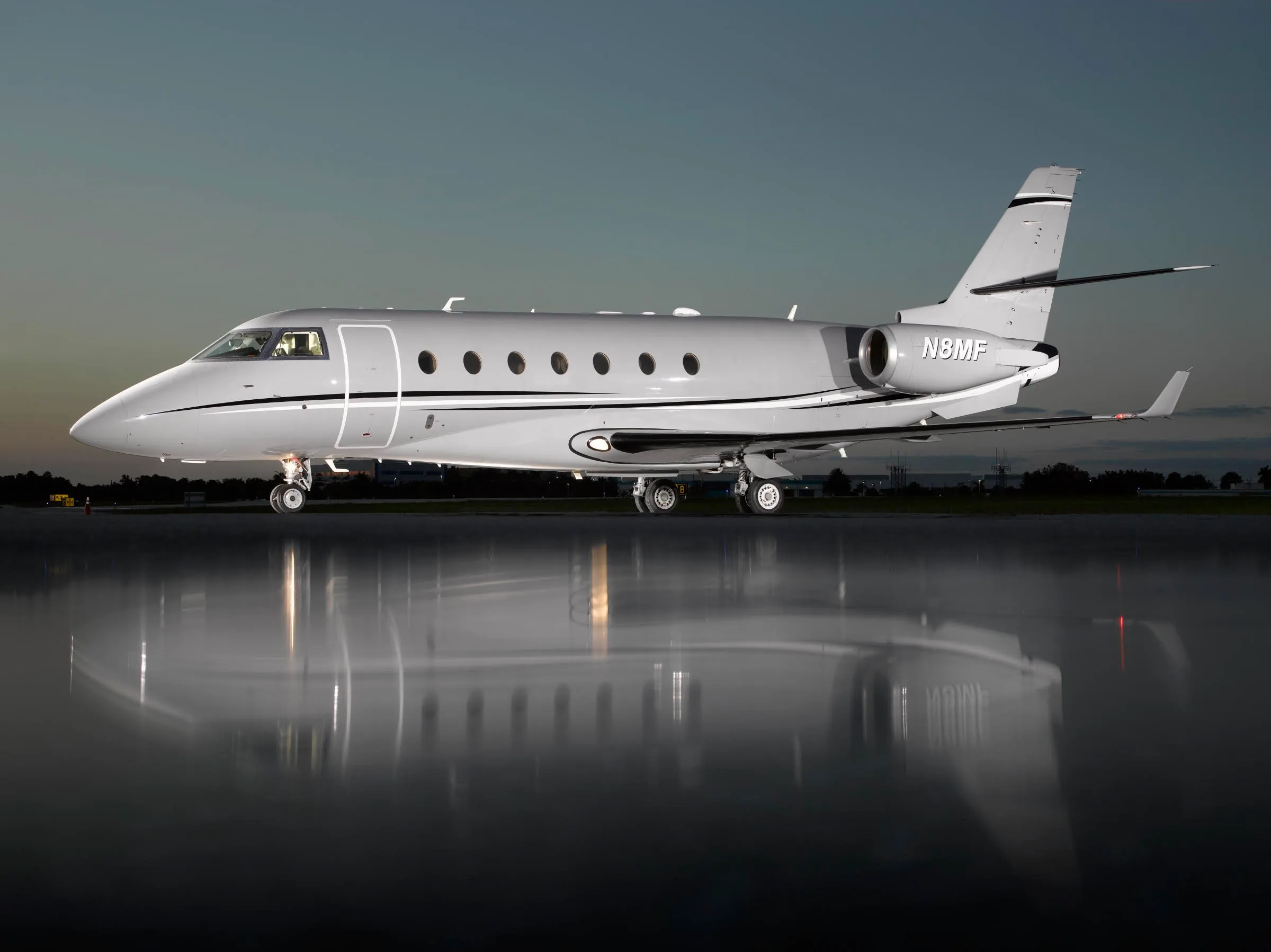 A sleek grey business jet, N8MF, is parked on a tarmac at dusk, with its reflection visible on the wet ground.
