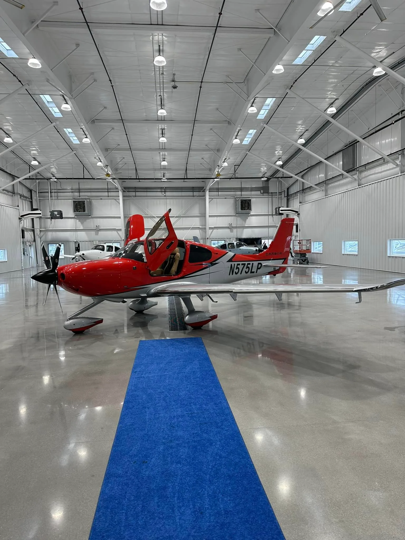 A red and silver Cirrus SR22 aircraft (N575LP) with its doors open, parked inside a brightly lit hangar on a blue carpet.
