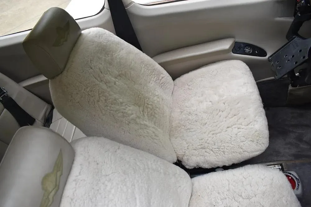 Interior view of the aircraft cabin showing seats with white sheepskin covers and a light-colored headrest with a logo.