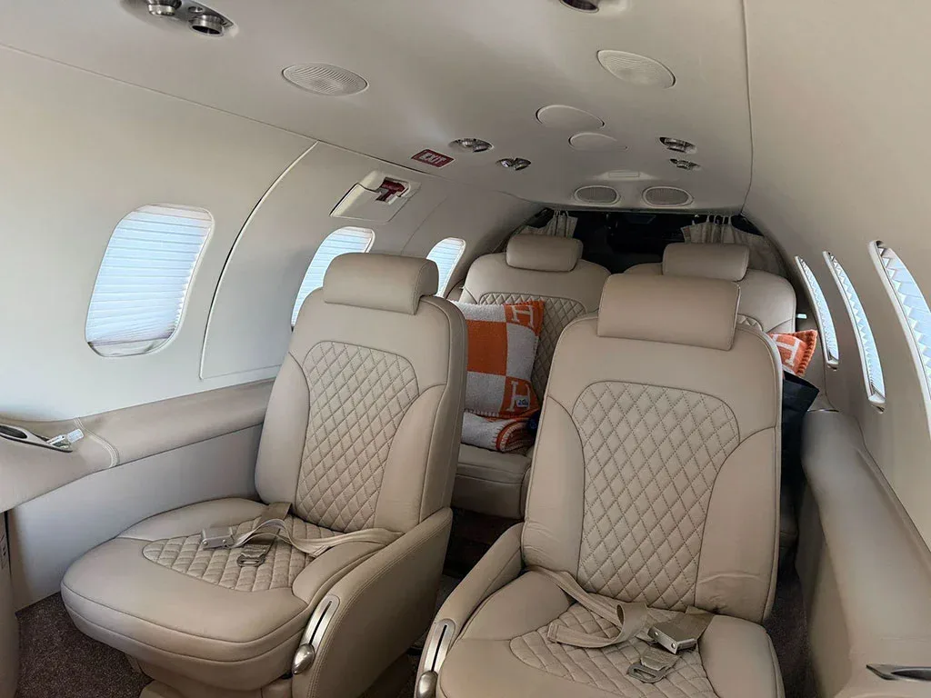 The aircraft cabin features light beige quilted leather seats and windows with blinds.