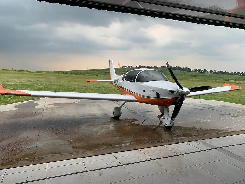 A white and orange light aircraft is parked on a wet concrete pad, partially under a hangar, with a green field and cloudy sky behind it.