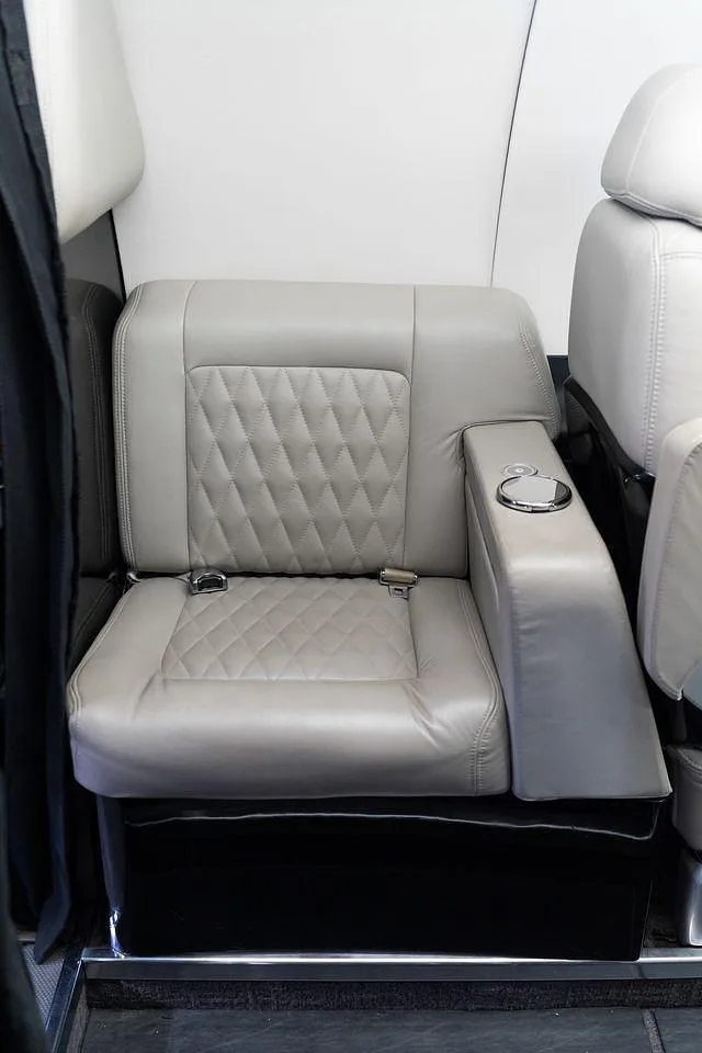 A light gray quilted leather passenger seat with an armrest and cup holder in the cabin of a business jet.
