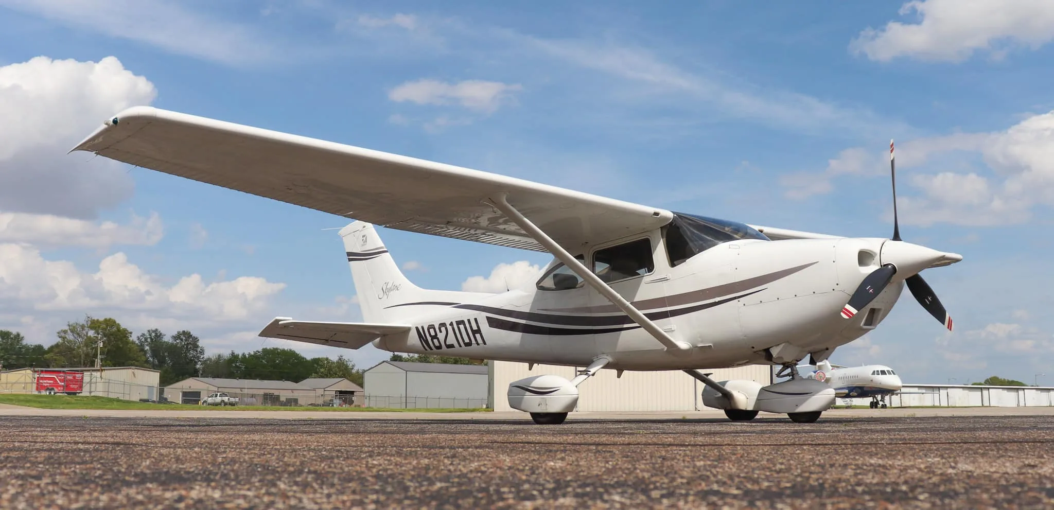 A low-angle front-right view of the white Cessna Skylane N821DH with dark grey and brown stripes, parked on an asphalt tarmac.